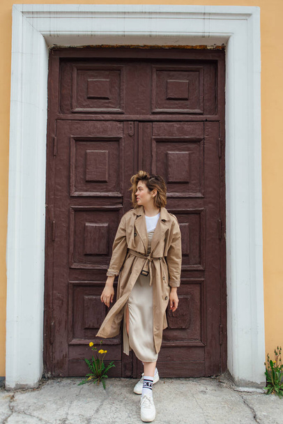 Young smiling millennial woman with wild hair dressed in an autumn coat posing near the door of an old building. - Fotografie, Obrázek