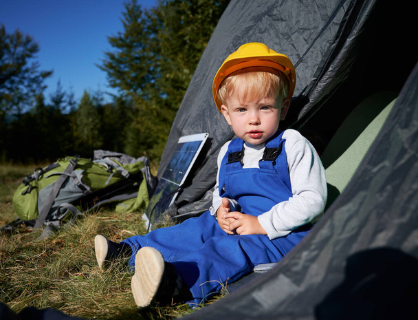 Cute male child in construction helmet looking at camera while sitting in tourist tent near portable solar panel and backpack. Adorable kid in work overalls resting in camp tent outdoors. - Photo, Image
