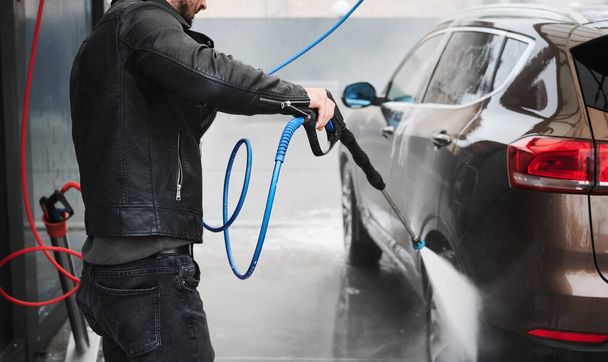 Cropped view of young man washing car on carwash station outdoor. Handsome driver cleaning automobile, using high pressure water. - 写真・画像