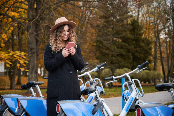 Közelről mosolygós fiatal nő göndör haj kalapban, és öltözött fekete elegáns kabát áll közel a modern kék kerékpár telefonnal. A városi parkban való kerékpározásra való felkészülés koncepciója. - Fotó, kép