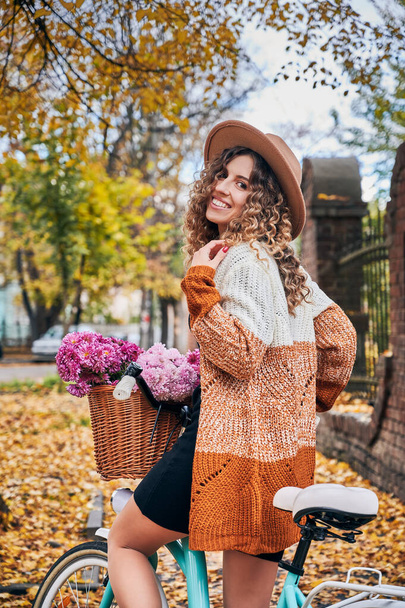 Close up of smiling young woman with curly hair in beige hat and dressed in stylish cardigan sitting on blue bicycle with basket beautiful flowers. Concept of walking on fresh air. - Photo, Image
