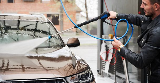 Cropped view of young man washing car on carwash station outdoor. Handsome worker cleaning automobile, using high pressure water. - Фото, зображення