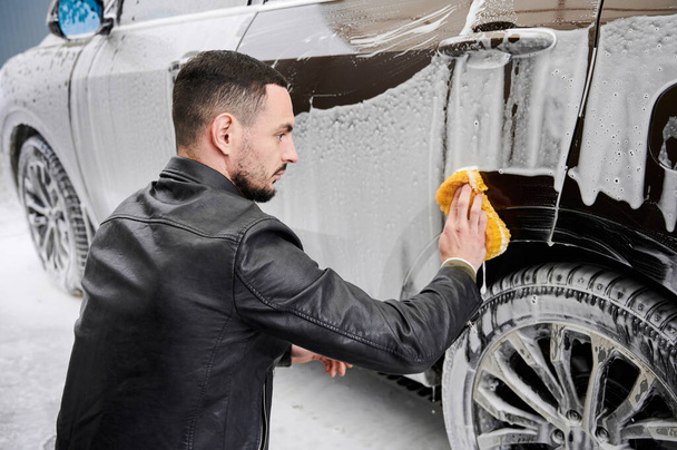 Hombre joven lavando coche en la estación de lavado de coches al aire libre. Conductor guapo de limpieza de automóviles, utilizando esponja y jabón. - Foto, imagen