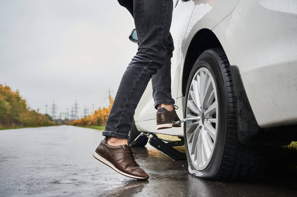 Close up of male auto mechanic standing on metal wrench while unscrewing lug nuts on car wheel. Young man using lug nut wrench while changing flat tire on the road. Concept of emergency road service. - Photo, Image