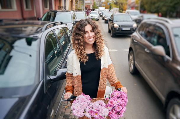 Front view of curly brunette travelling by vintage cycle with big basket of pink flowers. Pretty female wearing brown cardigan riding bike on street between autos. Concept of street style. - Foto, imagen