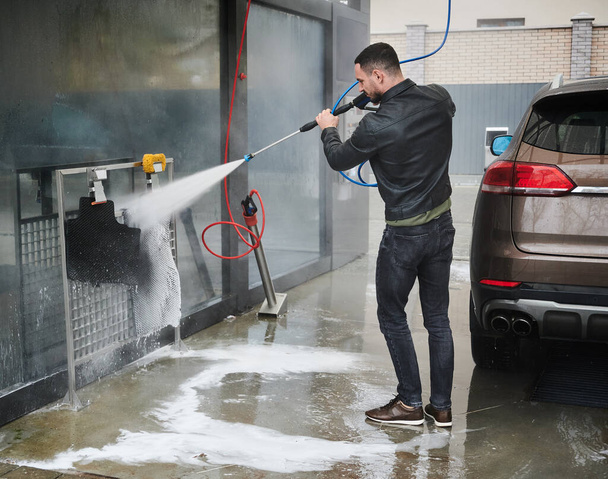 Self-service car wash station. Young male driver standing behind his car, holding in hands special equipment for washing vehicle. Man using strong jet of water pressure cleaning car mats. - Fotoğraf, Görsel