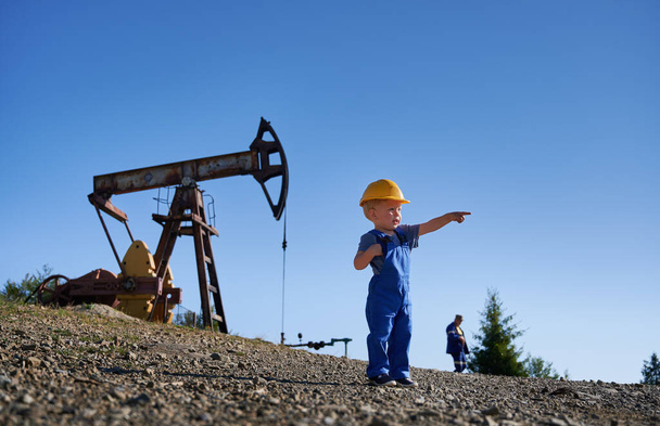Adorable child pointing finger at copy space while standing in oil field with petroleum pump jack and blue sky on background. Snapshot of oil filed with cute little boy and oil pump rocker-machine. - Photo, Image