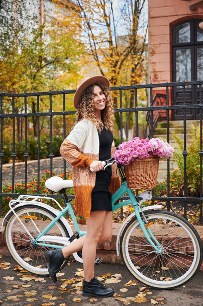 Vista lateral da mulher encaracolada viajando de bicicleta vintage rua abaixo. Mulher bonita vestindo chapéu marrom e casaco de lã está andando de bicicleta com grande cesta de camomilas rosa. Conceito de lazer. - Foto, Imagem