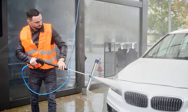 Jeune homme lavant sa voiture au lave-auto, vêtu d'un gilet orange. Beau travailleur nettoyage automobile blanche, en utilisant de l'eau à haute pression. - Photo, image