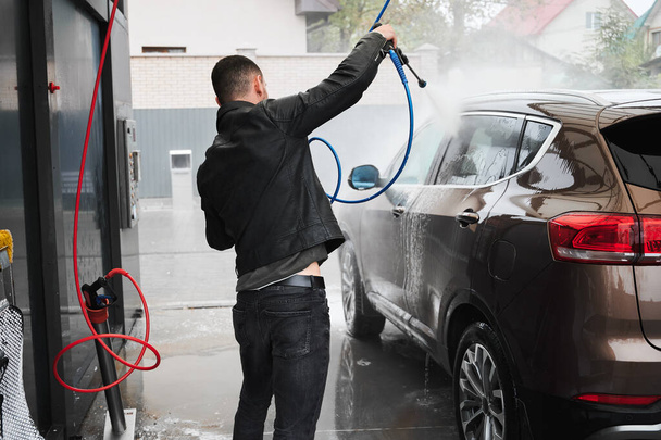 Rear view of luxury car owner who washing transport with running pressure water from gun. Automobile cleaning with professional equipment at self-service car wash. - Foto, imagen