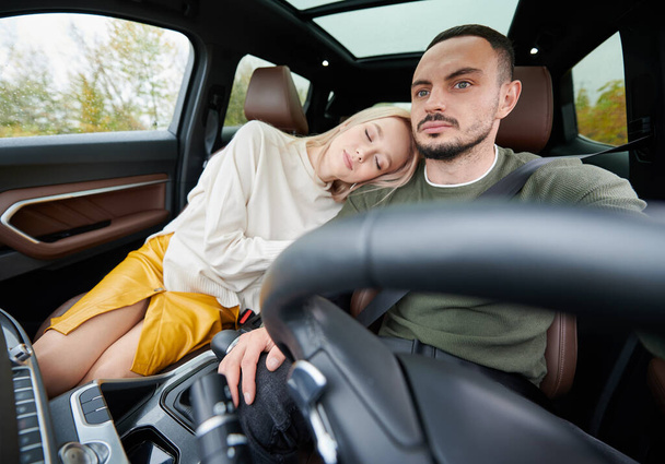 Happy man and woman traveling in car. Beautiful young couple sitting on the front passenger seats, handsome man driving comfortable automobile. Tired woman laying on guy's shoulder with closed eyes - Photo, image