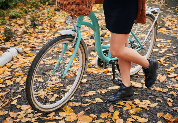 Detailní záběr na ženské nohy v elegantních botách a modré kolo na podzimní ulici. Mladá žena stojící na kole na asfaltu se žlutými listy, zatímco tráví čas venku. - Fotografie, Obrázek