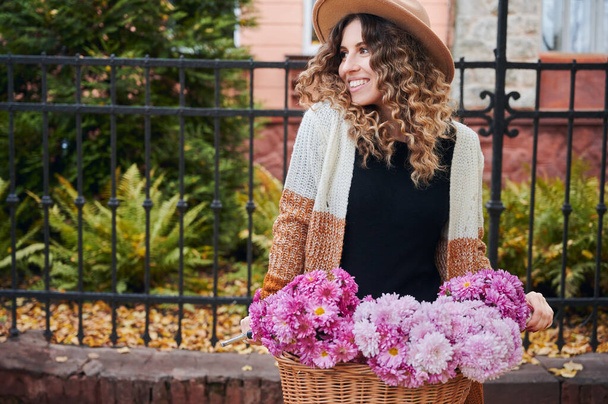 Side view of curly woman posing with bike near park fence. Smiling female wearing brown hat and cardigan is travelling by vintage cycle with big basket of pink camomiles. Concept of flowers. - Zdjęcie, obraz