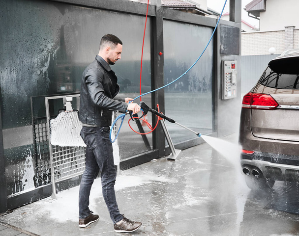 Молодий чоловік миє машину на парковці. Ручний робітник прибирає автомобіль, користуючись водою високого тиску.. - Фото, зображення