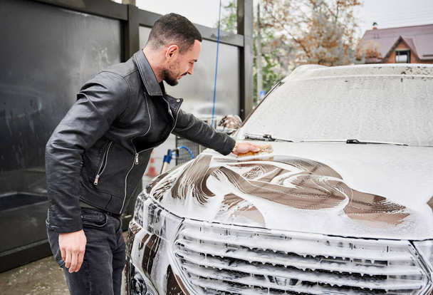 Araba yıkama istasyonunda araba yıkayan genç bir adam. Sünger ve sabun kullanarak araba temizleyen yakışıklı bir işçi.. - Fotoğraf, Görsel