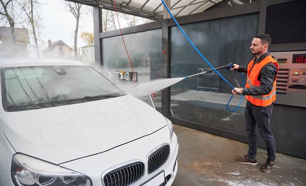 Young man washing car on carwash station outdoor, wearing orange vest. Handsome worker cleaning white automobile, using high pressure water. - Foto, afbeelding