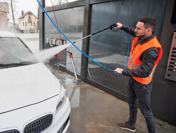 Young man washing white car on carwash station, wearing orange vest. Handsome worker cleaning automobile, using high pressure water. - Foto, afbeelding