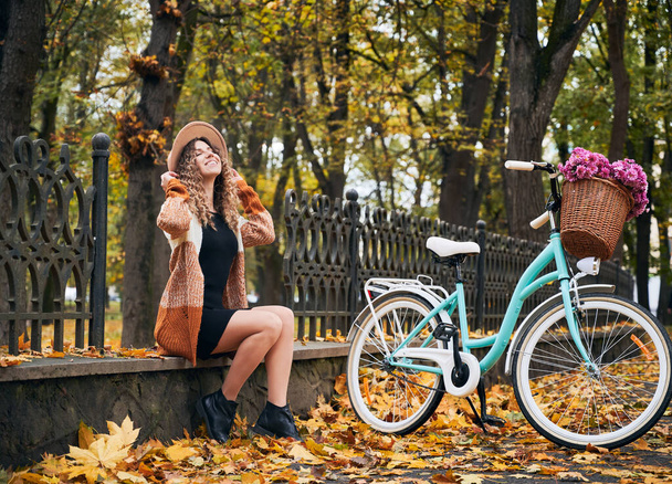 Side view of beautiful woman with retro bicycle is sitting on fence foundation in autumn park. Curly female is resting on settleand enjoy weather. Concept of joy of nature. - Photo, Image