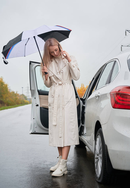 Beautiful woman standing on a road near her stopped white car with punctured car tire. Female driver calling car service for help, holding umbrella. - Photo, Image
