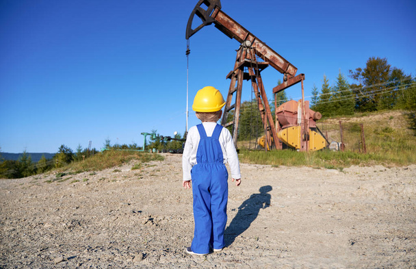 Πίσω όψη του χαριτωμένο παιδί στην εργασία φόρμες στέκεται στο έδαφος του τομέα του πετρελαίου με αντλία γρύλο και μπλε του ουρανού στο παρασκήνιο. Μικρό αγόρι στην κατασκευή κράνος κοιτάζοντας το πετρέλαιο αντλία rocker-μηχανή σε εξωτερικούς χώρους. - Φωτογραφία, εικόνα