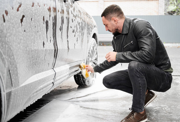 Young man washing car on carwash station outdoor. Handsome driver cleaning automobile, using sponge and soap. - Фото, изображение
