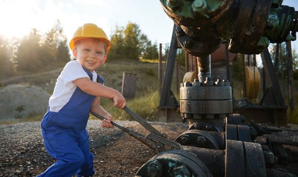 Cute cheerful child looking at camera and smiling while tightening bolt on pipe in oil field. Joyful kid in construction helmet using industrial wrench while repairing pump jack pipe on sunny day. - Photo, Image