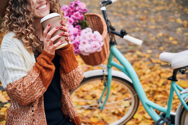 Close up de sorrir jovem mulher em cardigan elegante segurando xícara de café quente ou chá perto de bicicleta com flores frescas rosa bouquet. Conceito de descanso no ar fresco no parque urbano no outono. - Foto, Imagem