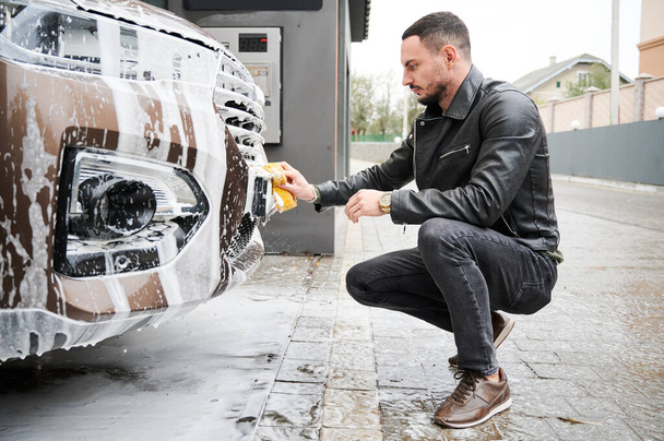 Hombre joven lavando coche en la estación de lavado de coches al aire libre. Hermoso trabajador de limpieza de automóviles, utilizando esponja y jabón. - Foto, imagen