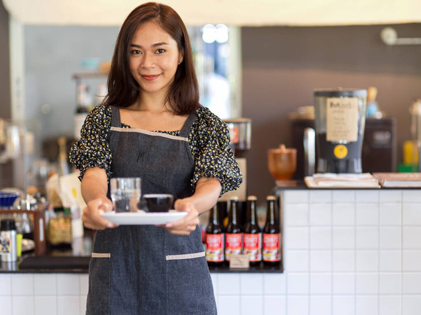 Portrait de la jeune femme confiante barista tenant un café avec plateau devant le comptoir du café - Photo, image