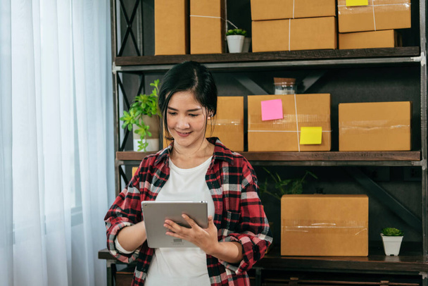 Asian woman startup small business at home office. Online seller entrepreneur young asian woman use tablet checking online order. Woman using tablet check stock goods supply delivery package shipping - Zdjęcie, obraz
