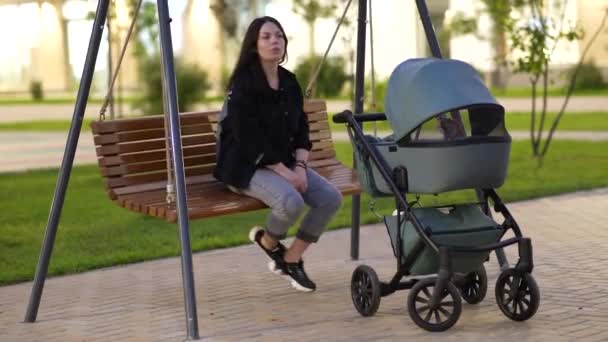 Maman avec une poussette est assise sur une balançoire dans la cour d'un immeuble résidentiel - Séquence, vidéo