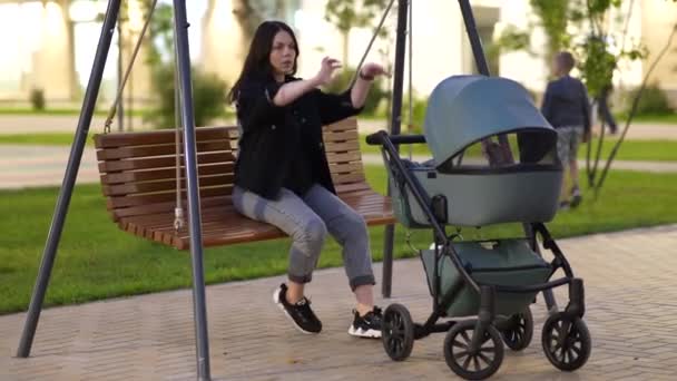 Maman avec une poussette est assise sur une balançoire dans la cour d'un immeuble résidentiel - Séquence, vidéo