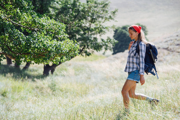 Side view of a woman with a read headband hiking in grassy hills. She's wearing small backpack, going downhill. - Foto, Imagen