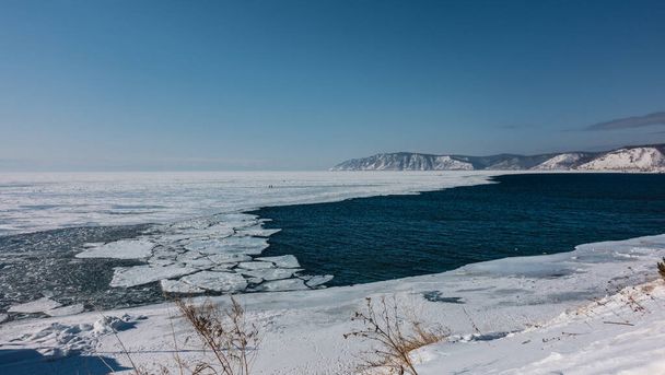 Aus einem zugefrorenen See fließt ein nicht zugefrorener Fluss. Blaues Wasser und weißes Eis. Eisschollen treiben auf der Oberfläche. Schnee und trockenes Gras am Ufer. Berge vor dem azurblauen Himmel. Angara - Foto, Bild