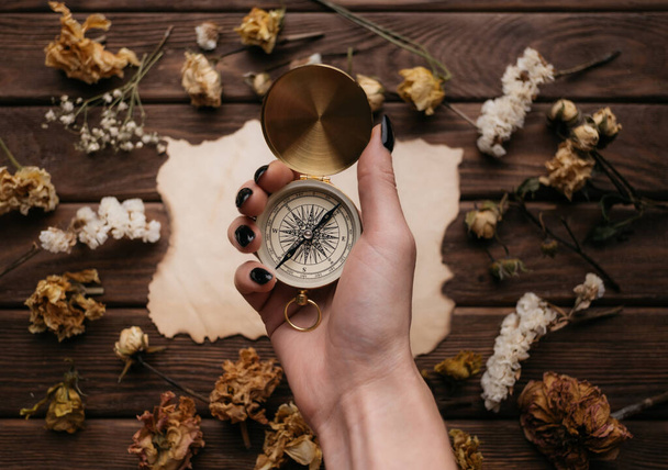 Vintage compass in female hands in the background a wooden table with dry flowers. View from above. - Фото, изображение