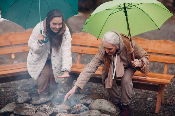 Donne che si rilassano al falò con ombrellone al campeggio glamping. Stile di vita vacanza moderno. - Foto, immagini