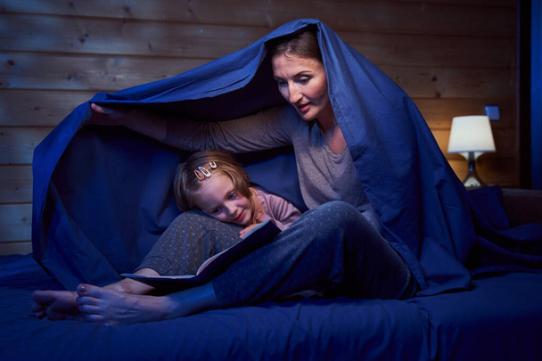 Female and child hiding under blanket while reading - Foto, Bild