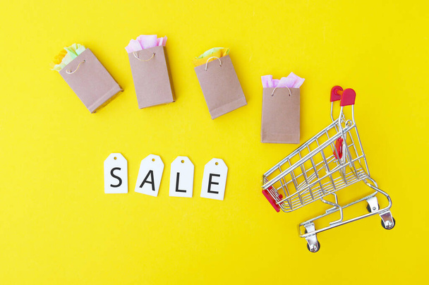 Shopping cart with paper bags and white labels with the word SALE on a yellow background. Shopping and discount concept. Black Friday. - Фото, изображение