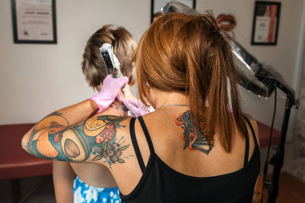 Lefthanders tatuador femenino con manga de tatuaje en guantes de color rosa hace un tatuaje a un cliente. Vista trasera. Día Internacional de los Lefthanders. - Foto, imagen