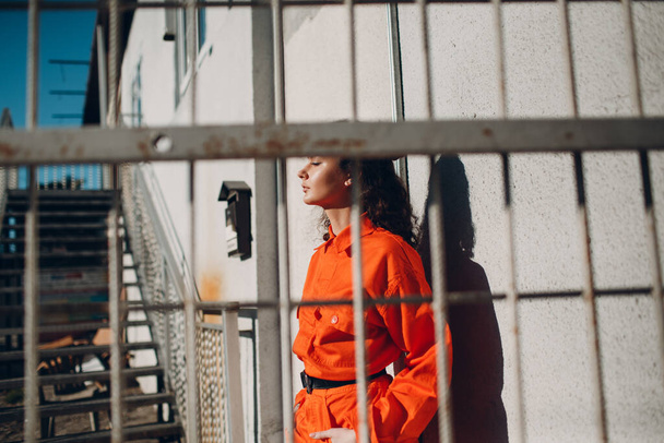 Jeune femme brune bouclée en costume orange derrière la cage. Femme en salopette colorée portrait. - Photo, image