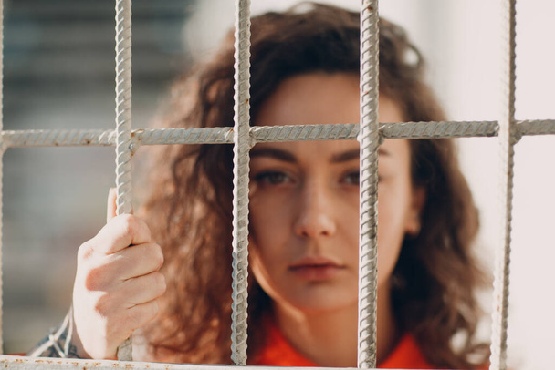 Young brunette curly woman in orange suit behind jail bars. Female in colorful overalls portrait. - Zdjęcie, obraz