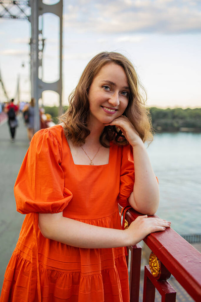 portret białej białej kobiety z czerwonymi włosami i niebieskimi oczami w pomarańczowej sukience stoi na moście. - Zdjęcie, obraz