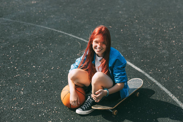 Una chica sonriente con una pelota de baloncesto está sentada en un monopatín en el campo de deportes - Foto, imagen