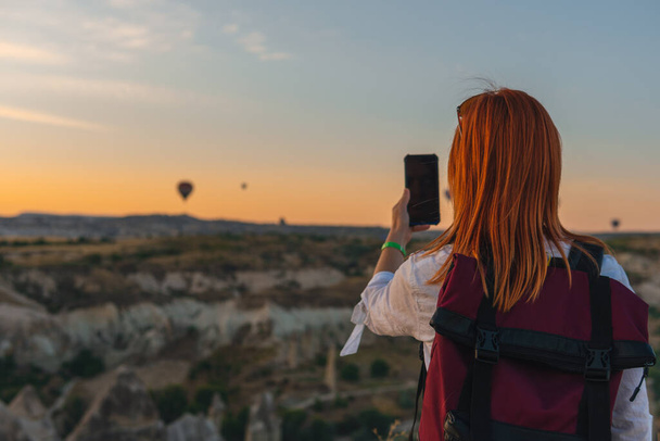 Girl with a backpack, takes a photo on a smartphone. valley with mountains and balloons at sunrise. Back view. Entertainment, tourism an vacation. Travel tour. Goreme, Cappadocia, Turkey. - Photo, Image
