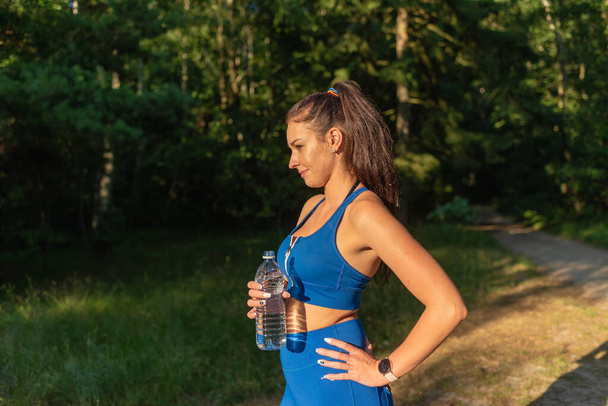 Девушка пьет воду из бутылки в лесу. Молодая женщина держит бутылку воды в лесу, портрет. позитивная женщина в спортивной форме отдыхает после бега, бега трусцой. в солнечный летний день - Фото, изображение