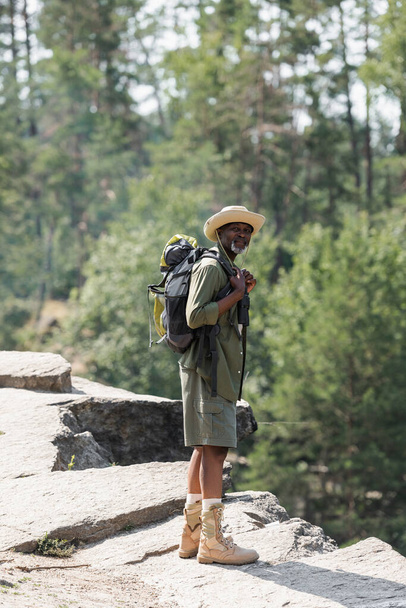 Senior african american hiker holding backpack on rock  - Photo, Image