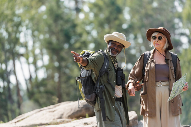 Sonriente excursionista afroamericano señalando con el dedo cerca de la esposa con mapa en el bosque  - Foto, imagen