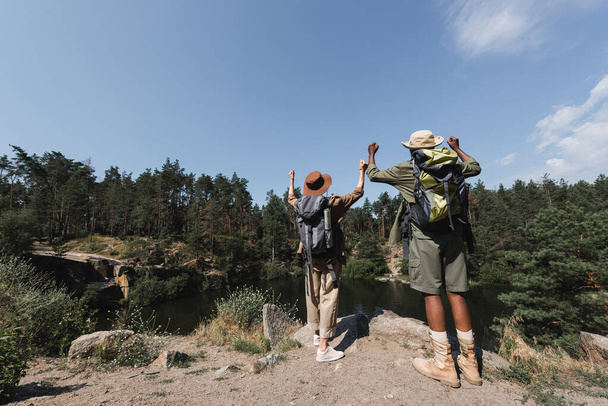 Back view of multiethnic hikers showing yes while standing on rock near lake  - Foto, immagini