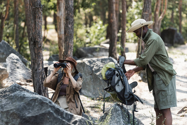Hombre afroamericano sosteniendo mochila cerca de esposa con prismáticos y piedras en el bosque  - Foto, Imagen