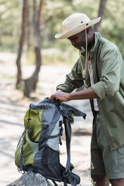 Старый африканский турист в шляпе держит рюкзак возле камня в лесу  - Фото, изображение
